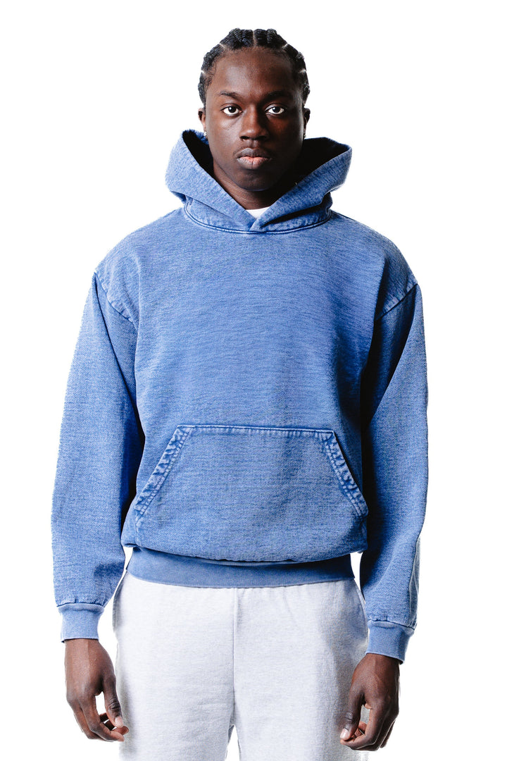 A person faces forward, wearing the Mineral Wash Heavy Fleece Hoodie in blue with the hood down and light gray pants against a white background.