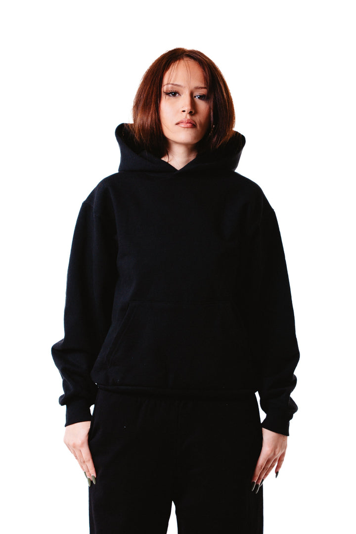 A person with straight, shoulder-length brown hair wears the Piece Dyed 14oz Heavy Fleece Hoodie and black pants, standing facing forward against a plain white background.