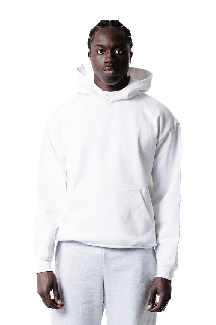 A person with braided hair faces forward, modeling the Piece Dyed 14oz Heavy Fleece Hoodie and light gray pants against a white background.